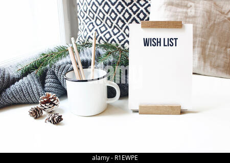 Winter desktop still life scene. Wish list card, board and wooden pencils in mug standing near window on white table background. Christmas concept, pi Stock Photo