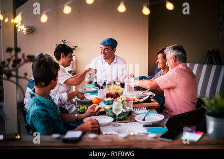 Dinner time in friendship with different ages people all together having fun and enjoying the night with smiles and happiness. Mobile phones defocused Stock Photo