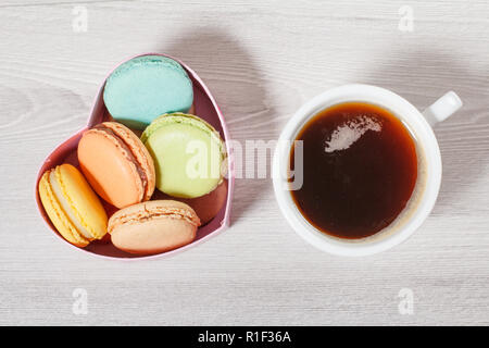Tasty macaroons cakes of different color in cardboard box in the shape of heart and cup of coffee on gray boards. Top view Stock Photo