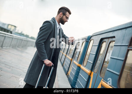 Business trip. Young businessman standing near railway with luggage checking time on watch waiting for train Stock Photo