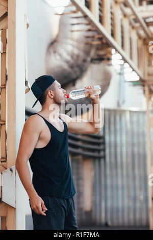 Young man drinking water from bottle. Sportsman resting after work out exercising in industrial city background Stock Photo