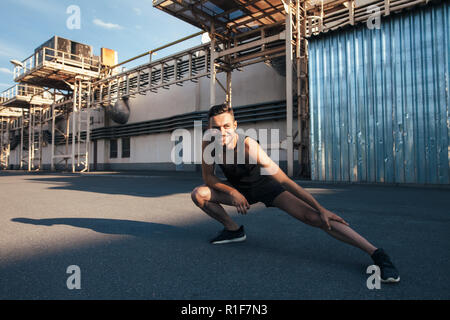 Smiling man doing warm up and stretching outdoor on in industrial city background. Fitness, training, healthy lifestyle Stock Photo