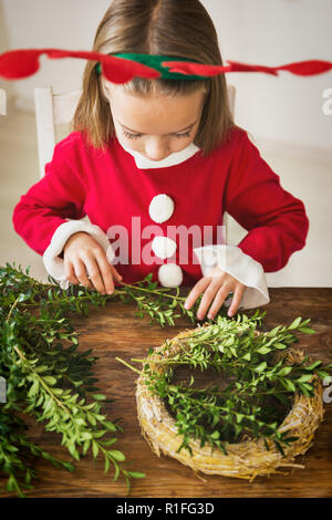 Cute preschooler girl dressed in reindeer costume wearing reindeer antlers making christmas wreath in living room. Christmas decoration family fun con Stock Photo