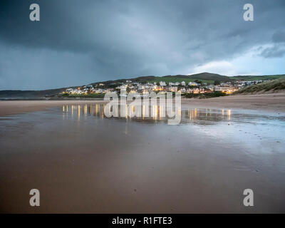 Woolacombe Beach, North Devon, UK 12th November 2018. Just before a rain storm threatening black rain clouds approach Woolacombe on the North Devon Coast. Lights from the seaside town are reflected in the beach at dusk after a bracing day of gusty wind, sun and showers. Credit: Julian Eales/Alamy Live News Stock Photo