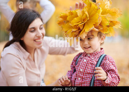 Mother puts yellow fallen leaves on son head. Happy family is in autumn city park. Children and parents. They posing, smiling, playing and having fun. Stock Photo