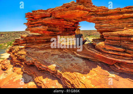 Blonde caucasian woman sitting inside the iconic rock arch in red sandstone of Nature's Window, Kalbarri National Park. Caucasian young girl in Western Australia looking australian outback landscape. Stock Photo