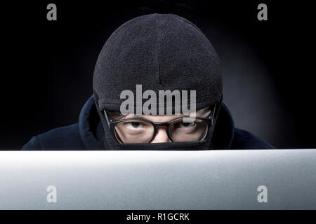 hacker in balaclava and glasses looks out from behind the laptop and looks into the camera Stock Photo