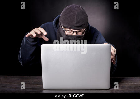 the cunning hacker in balaclava is sitting at the laptop and slyly looking at the camera Stock Photo