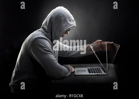 a hacker in a hood in profile sits at a laptop and starts to close it Stock Photo