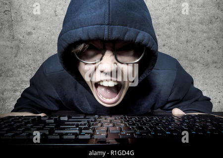hacker in the hood and glasses screaming furiously sitting in front of the keyboard Stock Photo