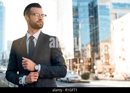 Close up profile portrait of a successful young bearded guy in suit and glasses. So stylish and nerdy. Outdoors on a sunny street, fixing his cuffs Stock Photo