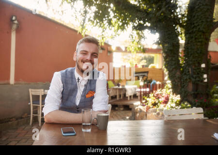 one young man, 20-29 years old, wearing hipster suit, sitting at table in cafe garden, looking to camera, smiling happy, looking to camera. Stock Photo