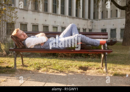 one young smiling man, 20-29 years old, wearing hipster retro suit, laying on bench in park. Stock Photo