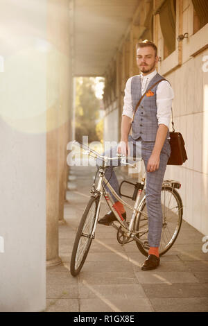 one young man, 20-29 years old,  wearing hipster suit, smart casual, posing sitting on a old city bike, sunny day, lens flare. building columns. Stock Photo