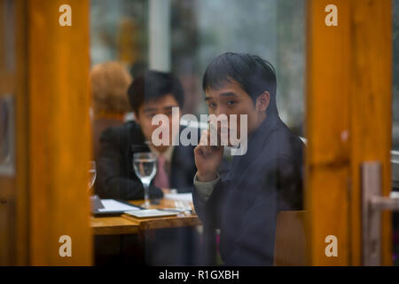 Young adult businessman sitting in a restaurant with a male colleague and using a cellphone. Stock Photo