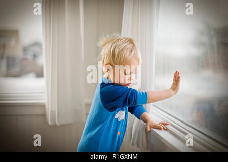 Young toddler pressing his hand against a window pane while standing inside a room. Stock Photo