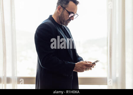 Mature businessman standing in hotel room using his cell phone. CEO on business trip texting on mobile phone in hotel room. Stock Photo