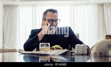 Happy mature businessman talking on mobile phone while sitting on dinning table in hotel room. Businessman having lunch and talking on phone in hotel Stock Photo