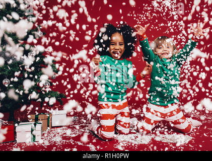 Two little girls in christmas costumes playing with artificial snow flakes. Kids having fun enjoying an artificial snowfall beside a christmas tree. Stock Photo