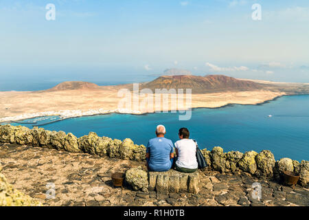 Two middle aged tourists sitting on a bench overlooking the small fishing and tourist village of Caleta del Sebo on the tiny island of La Graciosa nex Stock Photo