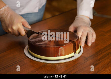 Prague mousse cake. Woman cuts off a piece of cake with a kitchen knife. Mirror glaze glitters deliciously. Modern cooking. Cooking in a pastry shop. Stock Photo
