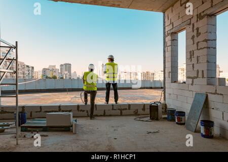 Two male builders at a construction site. Men looking into the distance to the panorama of the city. Building, development, teamwork and people concep Stock Photo