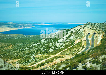 Two roads on a highway leading to tunnels in the Velebit mountain range among white rocks, green bushes and trees and azure blue sea water. Cloudless  Stock Photo