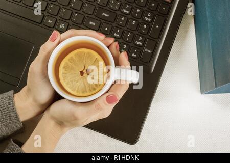 Top view of the keyboard on the female hands in a warm sweater with a cup of hot tea with lemon. Autumn and winter time, work in the office, at home. Stock Photo