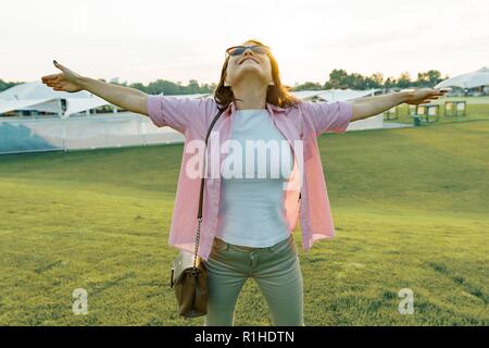 Middle-aged woman enjoys a vacation in summer recreation zone, female outdoors raising hands in sunlight park. Stock Photo