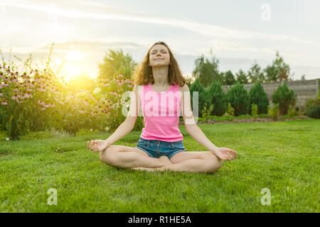 Young teenage girl practicing yoga, meditating on a summer sunset background, on green grass, lawn near the house Stock Photo