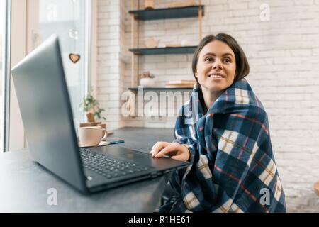 Autumn winter portrait of young smiling woman with laptop computer and cup of coffee in coffee shop, girl caught a cold covered with woolen plaid blan
