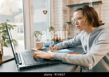 Portrait of young female student, schoolgirl in coffee shop with laptop computer and cup of coffee, girl is studying, writing in notebook. Stock Photo