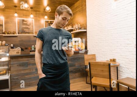 Young male cafe owner in an apron, with a mobile phone in the middle of a small coffee shop Stock Photo