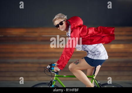 Photo on side of woman in red jacket and sunglasses riding bicycle in underpass against background of wooden benches Stock Photo