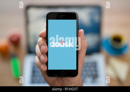 A man looks at his iPhone which displays the Zoosk logo, while sat at his computer desk (Editorial use only). Stock Photo
