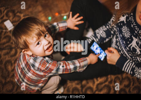 Baby boy sitting on the floor at home pointing at his mother’s smartphone Stock Photo