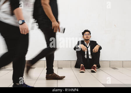 dowdy young asian businessman sitting at walkway thinking of his problem and look so depressed while people passing by ( motion blur ) Stock Photo