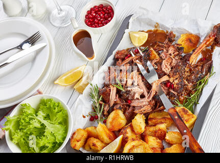 slow roast ultra tender shoulder of lamb pulled off the bones with crispy roast potatoes on a baking ray on a wooden table with glass of red wine and  Stock Photo