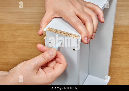 The furniture assembler puts a plastic pin into the hole on the surface of the wooden drawer with his thumb, with the other hand he holds the table, r Stock Photo