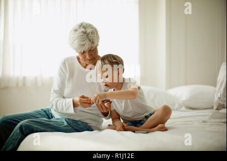 Mature woman in her pajamas sits on a bed with her young grandson and shows him a cell phone. Stock Photo