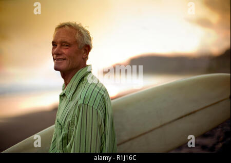 Mature man smiles as he stands on a beach carrying a surfboard and poses for a portrait. Stock Photo
