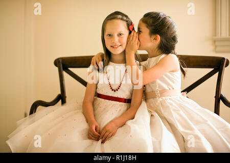 Young girls in white party dresses sit on wooden bench and whisper secrets as they pose for a portrait. Stock Photo