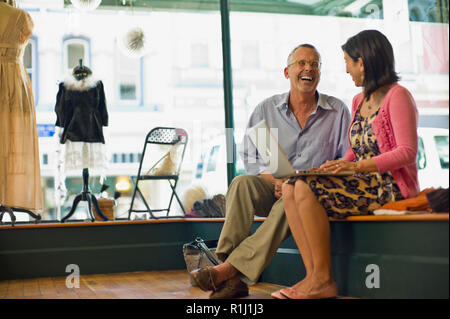 Laughing mature man sitting next to her wife as she uses her laptop inside a clothing store. Stock Photo