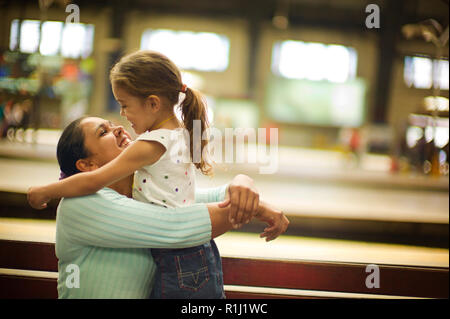 Mother and daughter smile at each other as the young girl stands on a bench and hugs her sitting mother. Stock Photo