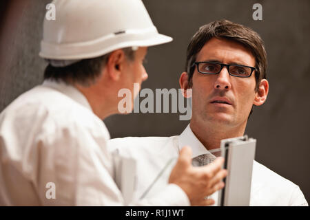 Businessman wearing eyeglasses looks concerned as a a male colleague in a hardhat talks to him. Stock Photo