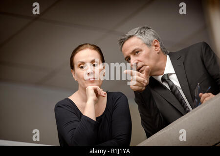 Serious businessman and a female colleague lean on the edge of an interior office balconey and look down as the businessman points to something below. Stock Photo