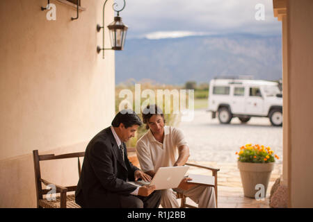 Two mid-adult men looking at a laptop while seated next to each other on a porch. Stock Photo