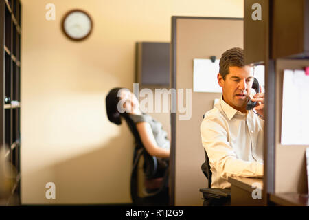 Two business people sit in office cubicles, one talking on a phone. Stock Photo