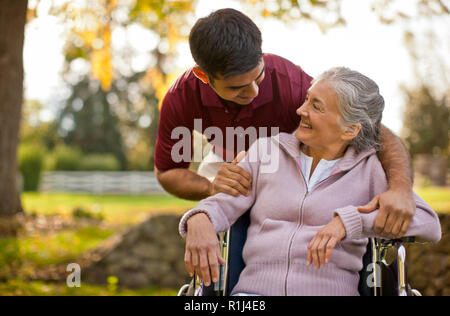 Smiling senior woman being comforted by a male nurse while sitting in a wheelchair inside a park. Stock Photo