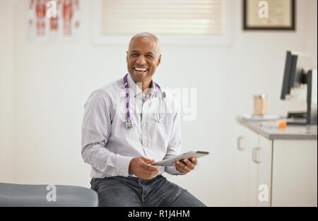 Portrait of a laughing male doctor holding a digital tablet while sitting inside his office. Stock Photo